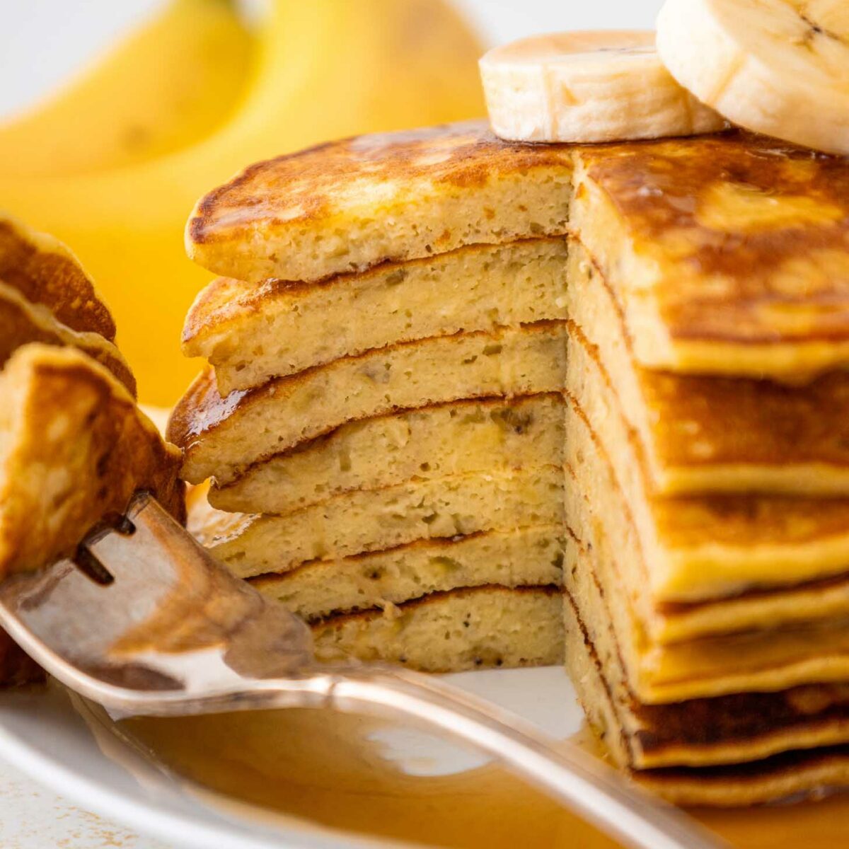 A stack of sliced banana pancakes topped with banana slices sits on a white plate, with syrup drizzled over the pancakes. A fork holds a piece of pancake, and bananas are visible in the background.