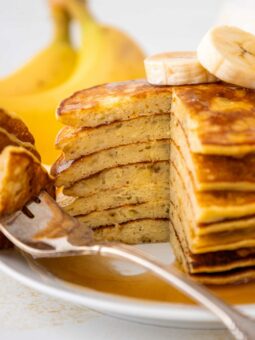A stack of sliced banana pancakes topped with banana slices sits on a white plate, with syrup drizzled over the pancakes. A fork holds a piece of pancake, and bananas are visible in the background.
