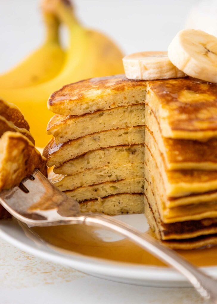 A stack of sliced banana pancakes topped with banana slices sits on a white plate, with syrup drizzled over the pancakes. A fork holds a piece of pancake, and bananas are visible in the background.