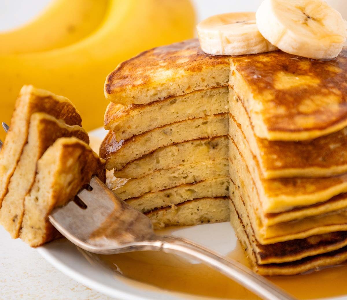 A stack of sliced banana pancakes topped with banana slices sits on a white plate, with a fork holding a cut piece. A whole banana is visible in the background. Syrup is drizzled on the pancakes.