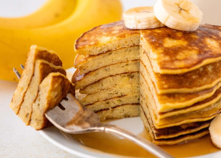 A stack of sliced banana pancakes topped with banana slices sits on a white plate, with a fork holding a cut piece. A whole banana is visible in the background. Syrup is drizzled on the pancakes.