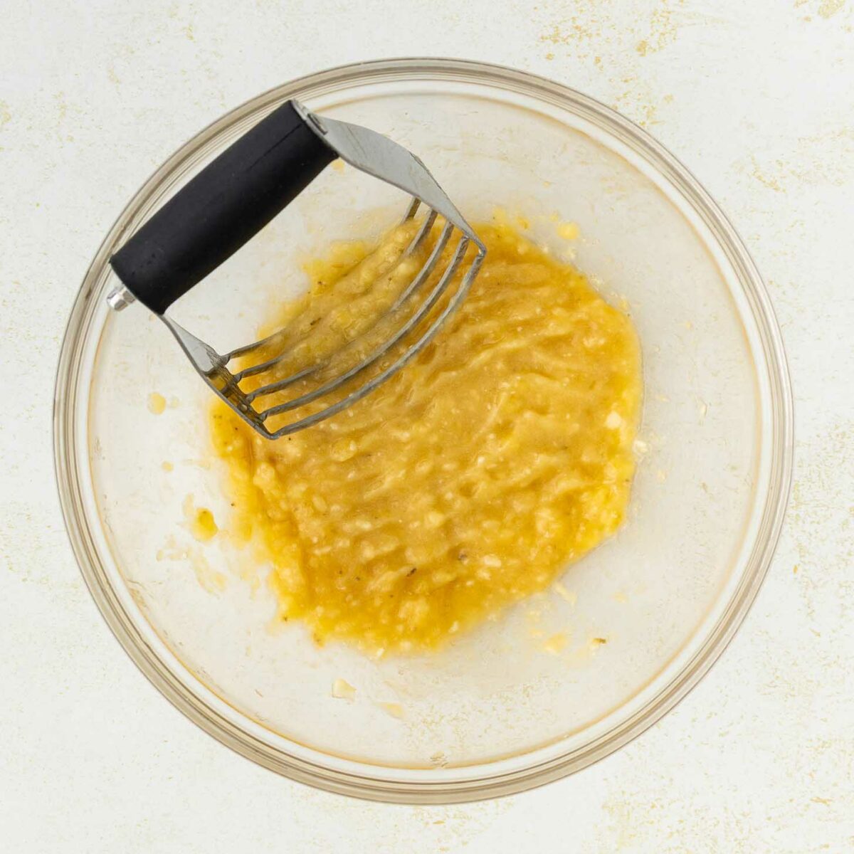 A glass bowl filled with mashed bananas and a metal potato masher, resting on a light-colored surface.