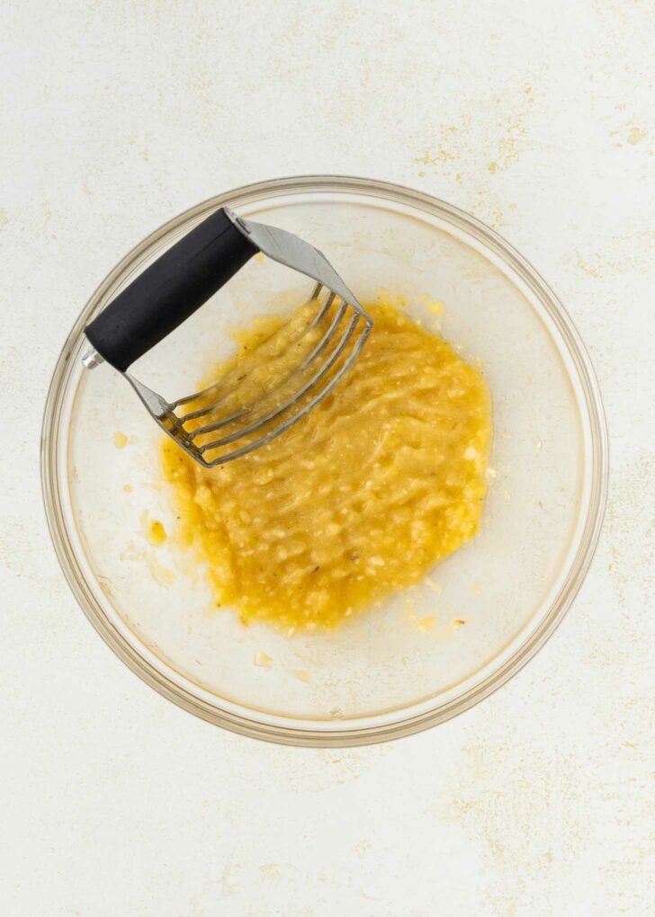 A glass bowl filled with mashed bananas and a metal potato masher, resting on a light-colored surface.
