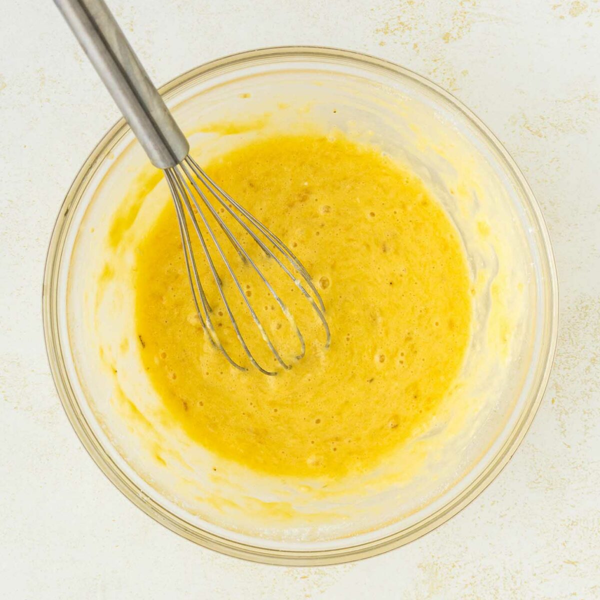 A metal whisk rests in a glass bowl filled with a yellow, foamy batter mixture on a light-colored surface.