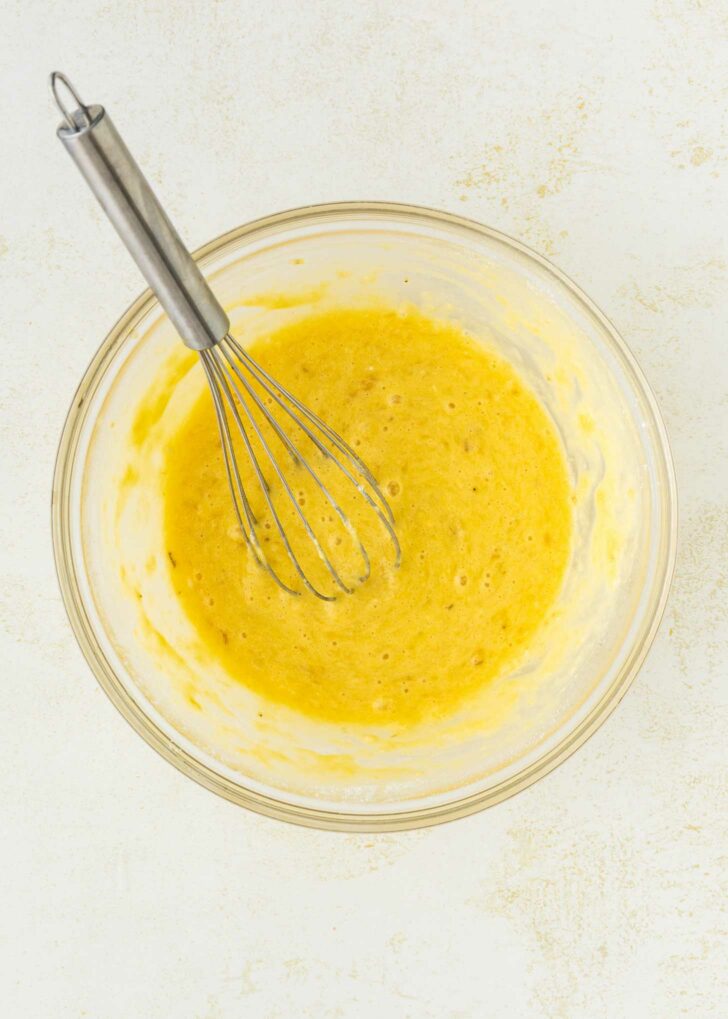 A metal whisk rests in a glass bowl filled with a yellow, foamy batter mixture on a light-colored surface.