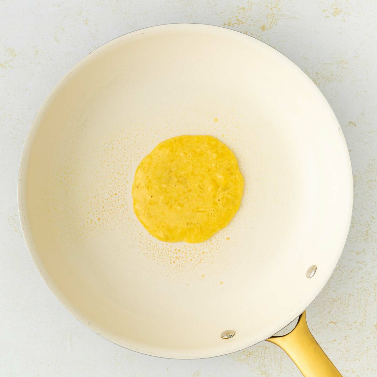 A single pancake is cooking in the center of a light-colored nonstick frying pan with a gold handle, set against a white background.