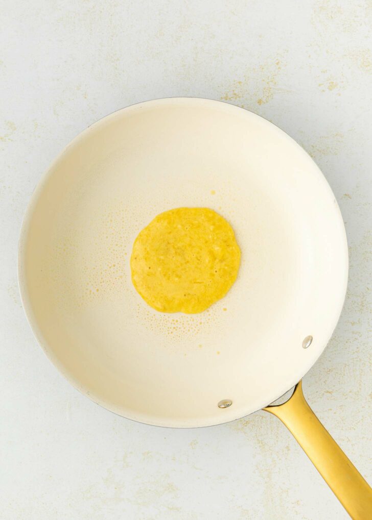 A single pancake is cooking in the center of a light-colored nonstick frying pan with a gold handle, set against a white background.