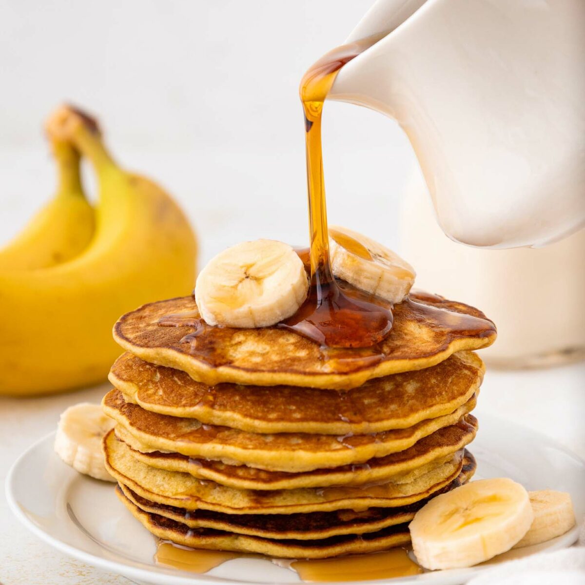A stack of banana pancakes topped with banana slices is on a white plate. Syrup is being poured from a white pitcher onto the pancakes. A banana and cut banana slices are nearby.
