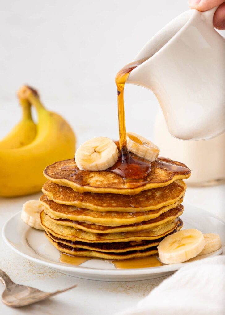 A stack of banana pancakes topped with banana slices is on a white plate. Syrup is being poured from a white pitcher onto the pancakes. A banana and cut banana slices are nearby.