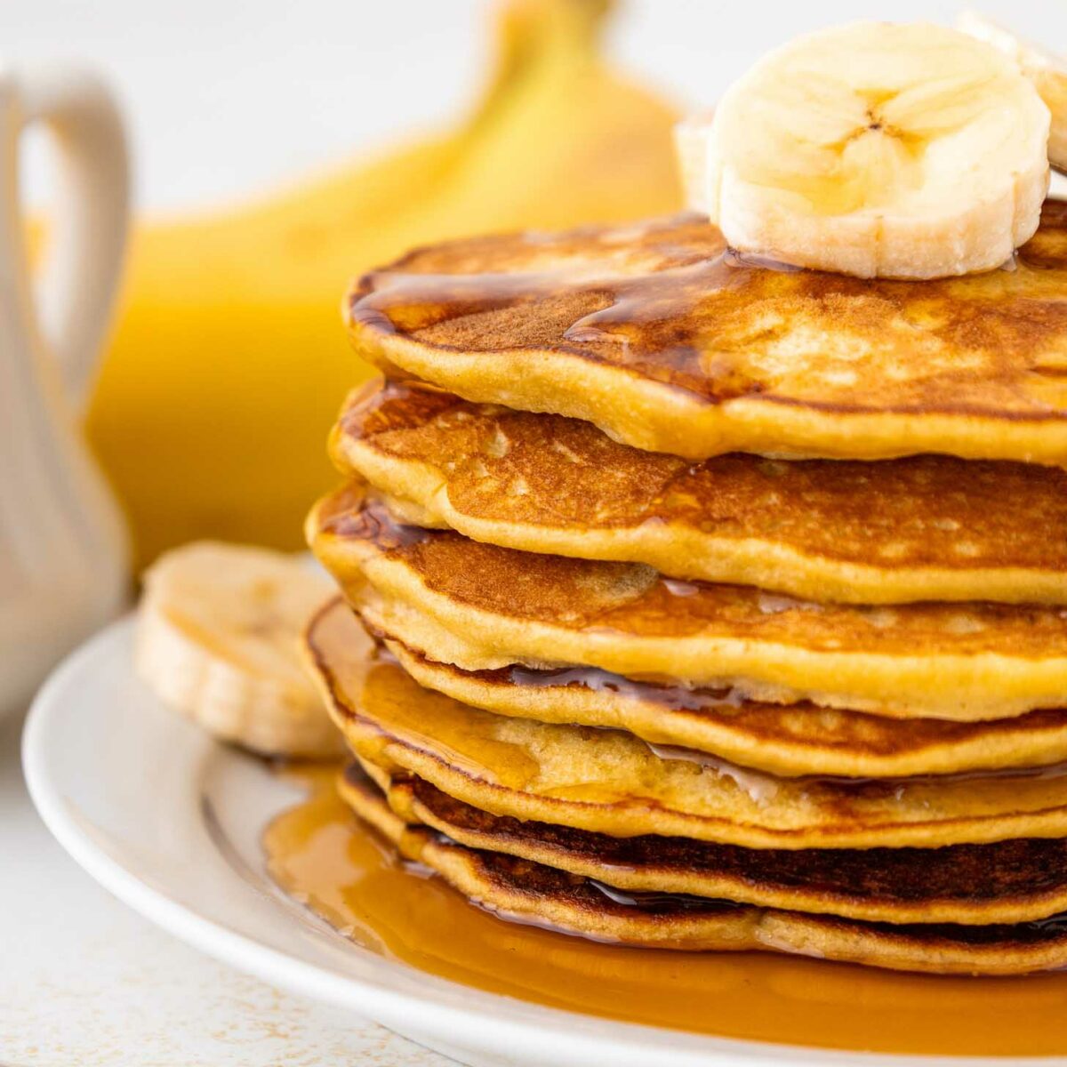 A stack of golden pancakes topped with banana slices and drizzled with syrup, served on a white plate with a banana and a syrup pitcher in the background.
