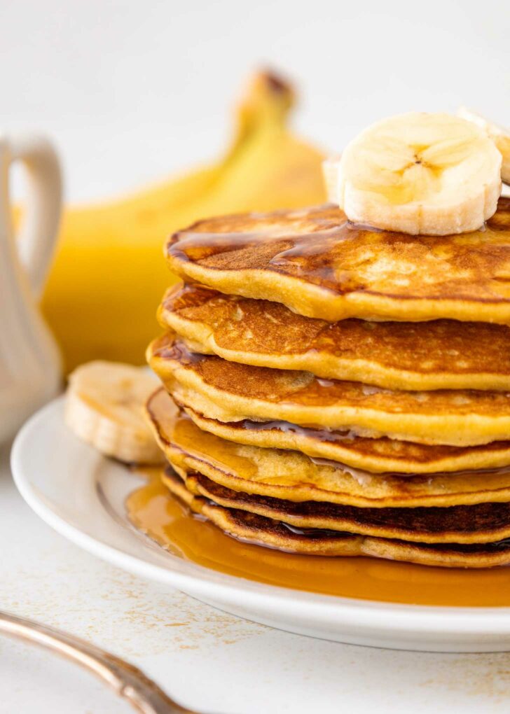 A stack of golden pancakes topped with banana slices and drizzled with syrup, served on a white plate with a banana and a syrup pitcher in the background.