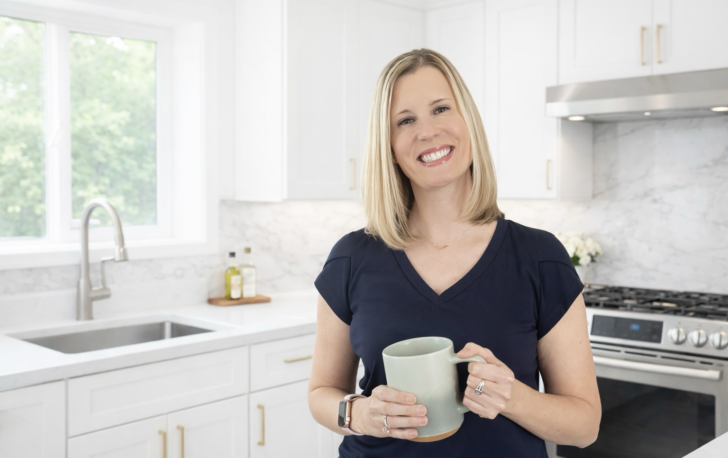 A smiling woman with blonde hair, wearing a navy blue shirt, stands in a bright, modern kitchen holding a large light green mug. White cabinets, a sink, and a stove are visible in the background.