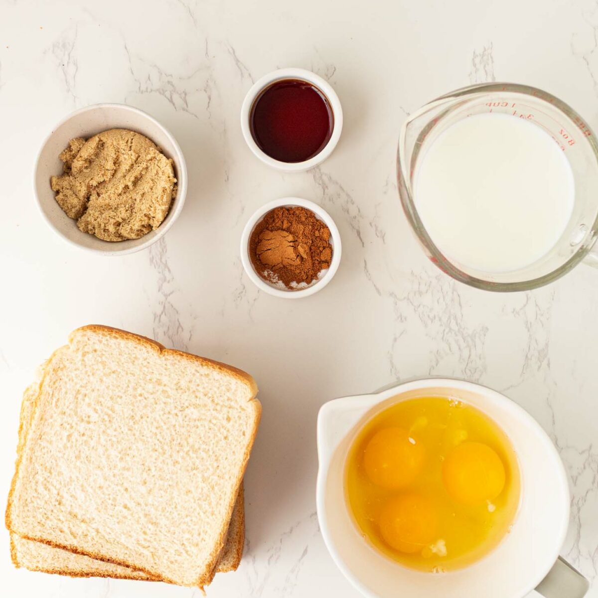 A flat lay of ingredients on a white marble surface: three slices of white bread, a bowl with three eggs, a measuring cup of milk, and small bowls of brown sugar, vanilla extract, and cinnamon.