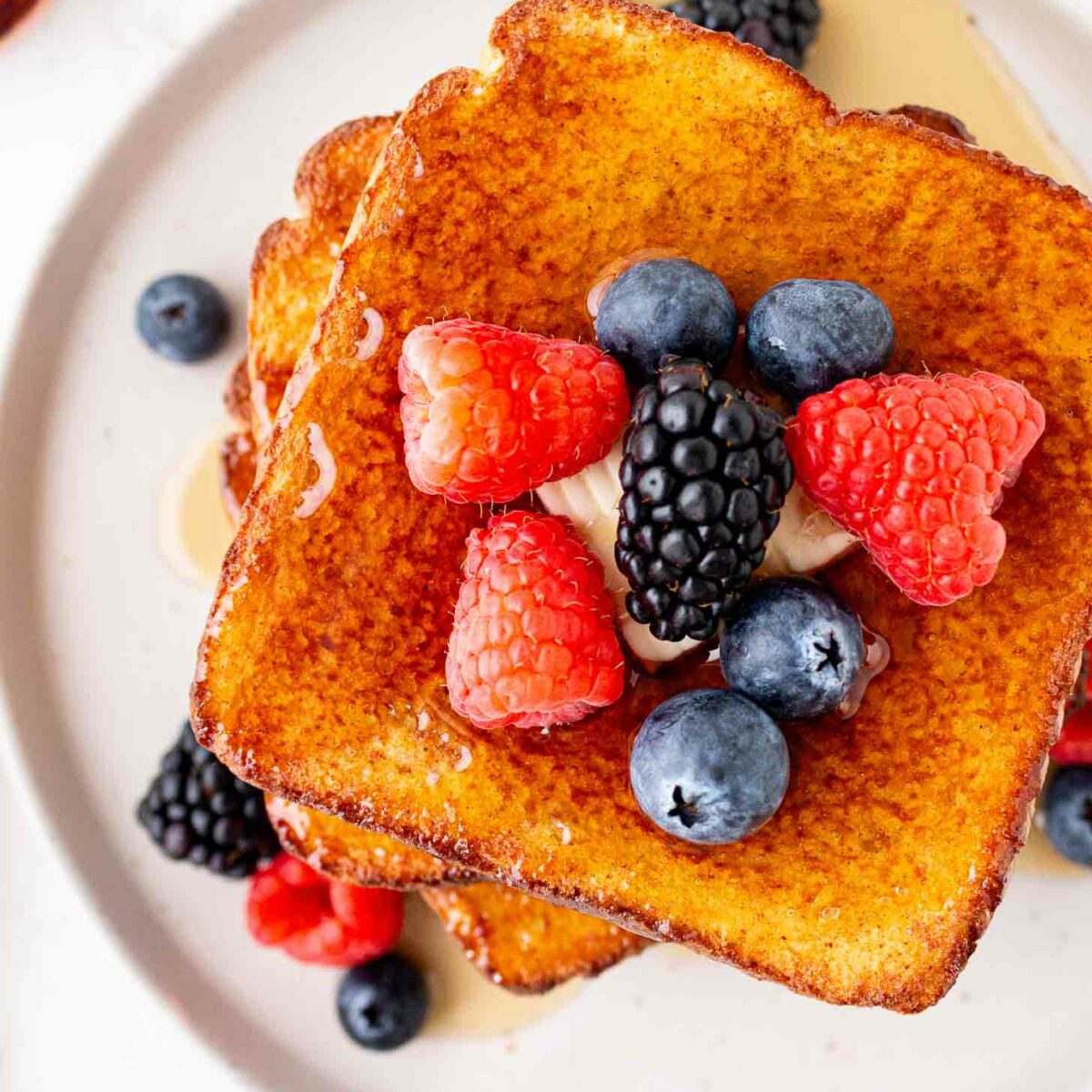 A stack of golden-brown French toast topped with fresh raspberries, blackberries, blueberries, and a dollop of cream or butter, served on a white plate with syrup drizzled around.