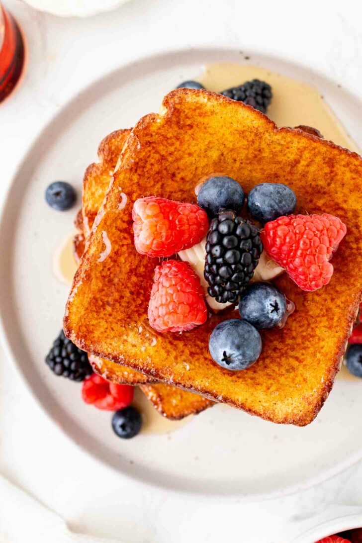 A stack of golden-brown French toast topped with fresh raspberries, blackberries, blueberries, and a dollop of cream or butter, served on a white plate with syrup drizzled around.