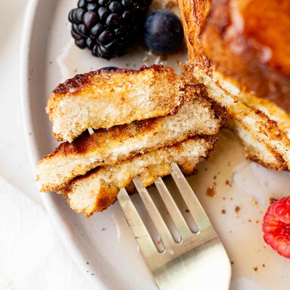 A close-up of a fork holding a cut piece of French toast on a plate, with syrup drizzled on top and fresh blackberries, blueberries, and a raspberry beside it.