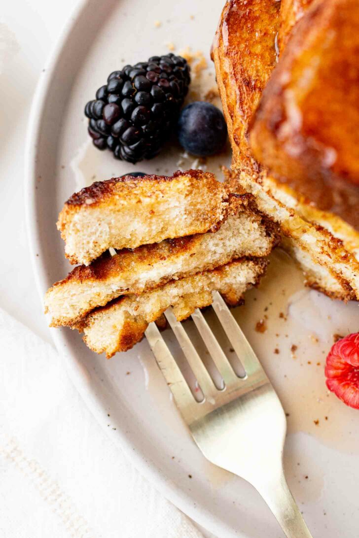 A close-up of a fork holding a cut piece of French toast on a plate, with syrup drizzled on top and fresh blackberries, blueberries, and a raspberry beside it.