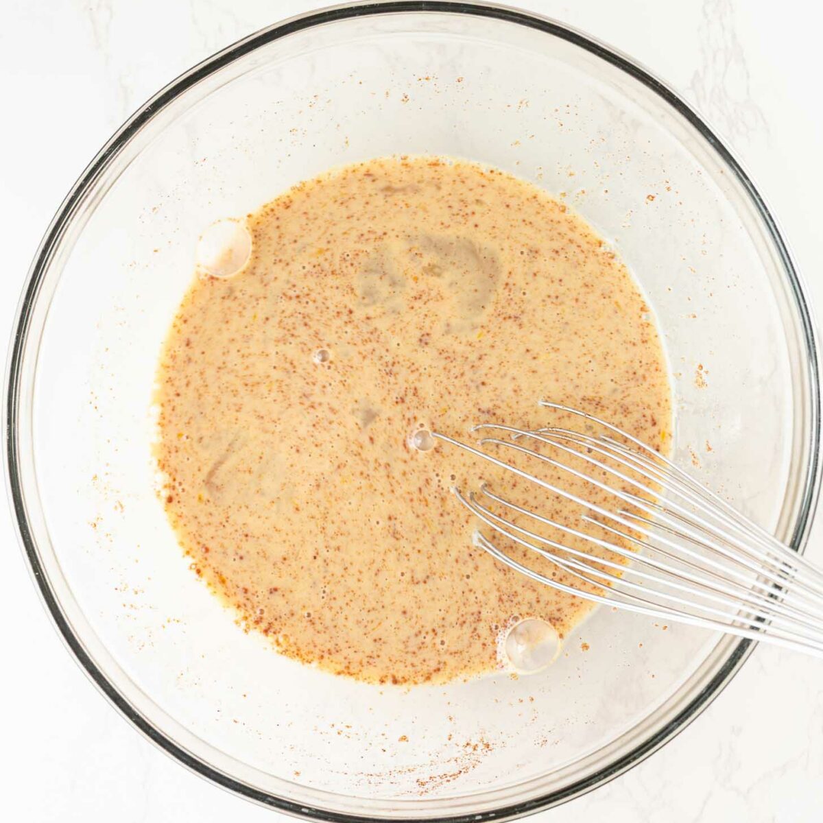 A glass mixing bowl filled with a light brown, speckled liquid mixture and a metal whisk resting inside on a white marble surface.