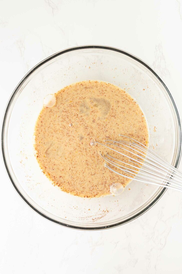 A glass mixing bowl filled with a light brown, speckled liquid mixture and a metal whisk resting inside on a white marble surface.