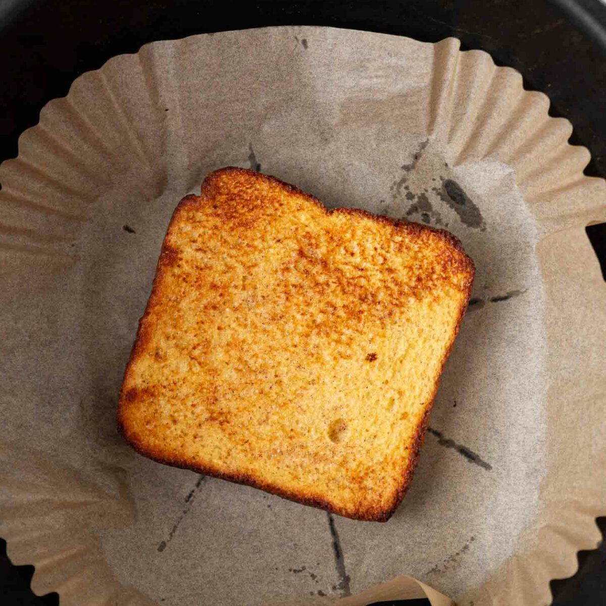 A single slice of toasted bread with golden brown edges sits on parchment paper inside a round black air fryer basket.