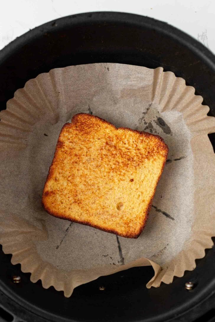 A single slice of toasted bread with golden brown edges sits on parchment paper inside a round black air fryer basket.