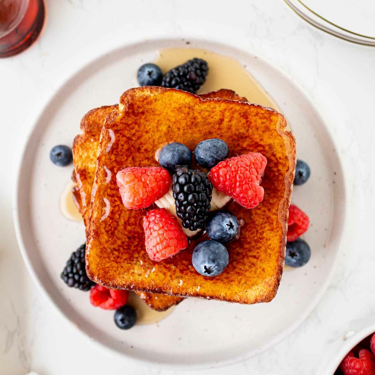 Two slices of French toast topped with fresh raspberries, blueberries, and blackberries, drizzled with syrup, served on a white plate. Butter, syrup, and extra berries are visible around the plate.