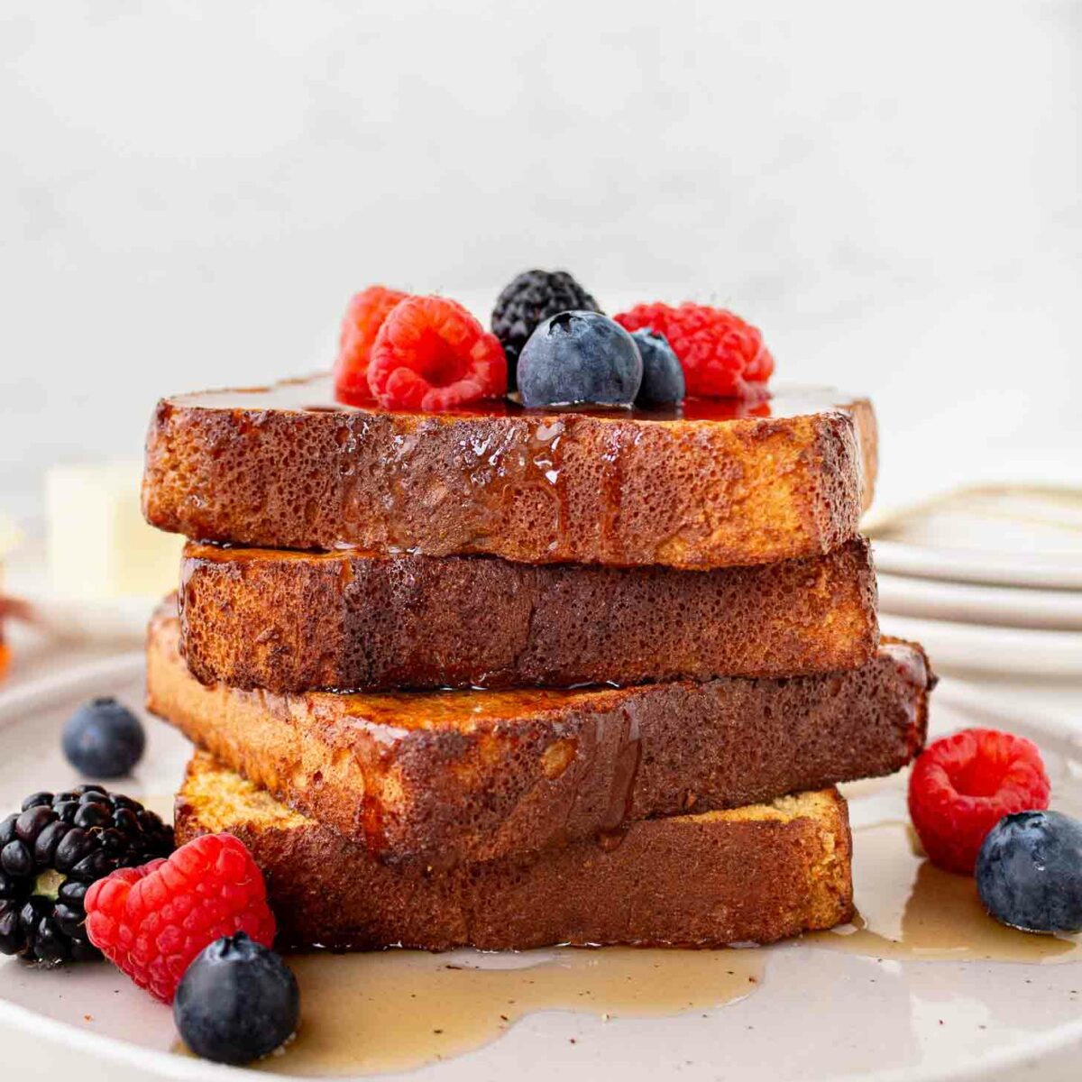 A stack of French toast slices topped with fresh raspberries, blueberries, and blackberries, drizzled with syrup, served on a white plate.