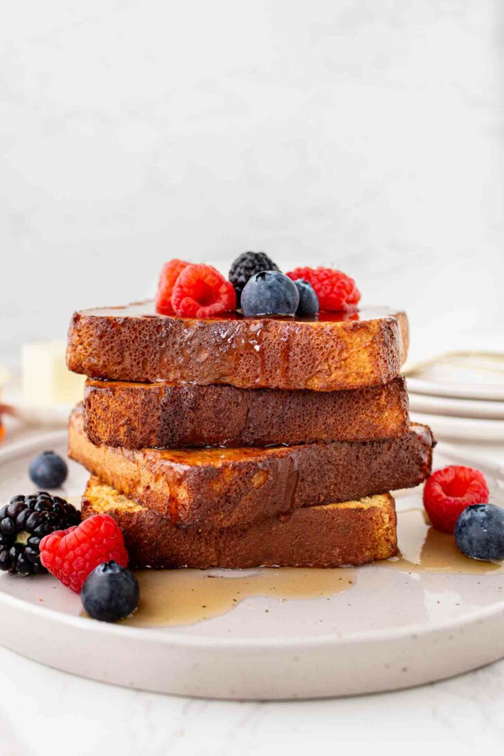 A stack of French toast slices topped with fresh raspberries, blueberries, and blackberries, drizzled with syrup, served on a white plate.