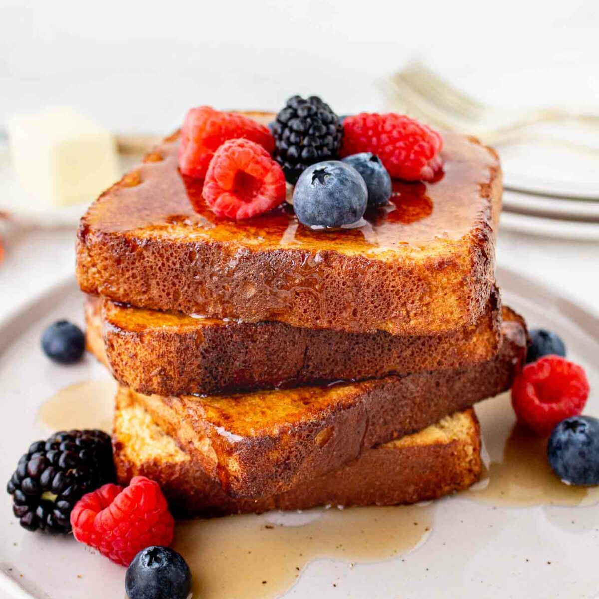 A stack of three thick slices of French toast topped with syrup, fresh raspberries, blueberries, and a blackberry, served on a light-colored plate with extra berries around the toast.