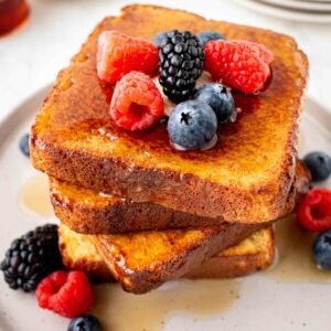 A stack of French toast topped with fresh raspberries, blackberries, and blueberries, drizzled with syrup, served on a white plate with extra berries on the side.