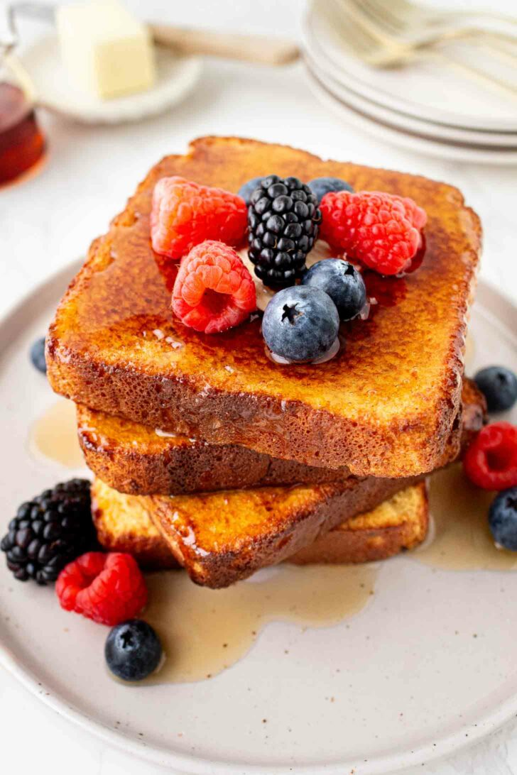 A stack of French toast topped with fresh raspberries, blackberries, and blueberries, drizzled with syrup, served on a white plate with extra berries on the side.