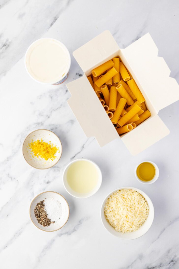 A flat lay of dry rigatoni pasta in an open box, grated cheese, ricotta, olive oil, lemon zest, black pepper, and salt in small bowls on a white marble surface.