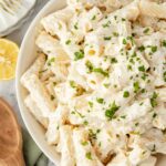 A bowl of rigatoni pasta covered in creamy white sauce, garnished with chopped parsley. A wooden spoon, lemon half, and forks are nearby on a marble surface.