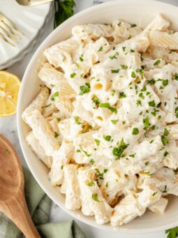 A bowl of rigatoni pasta covered in creamy white sauce, garnished with chopped parsley. A wooden spoon, lemon half, and forks are nearby on a marble surface.