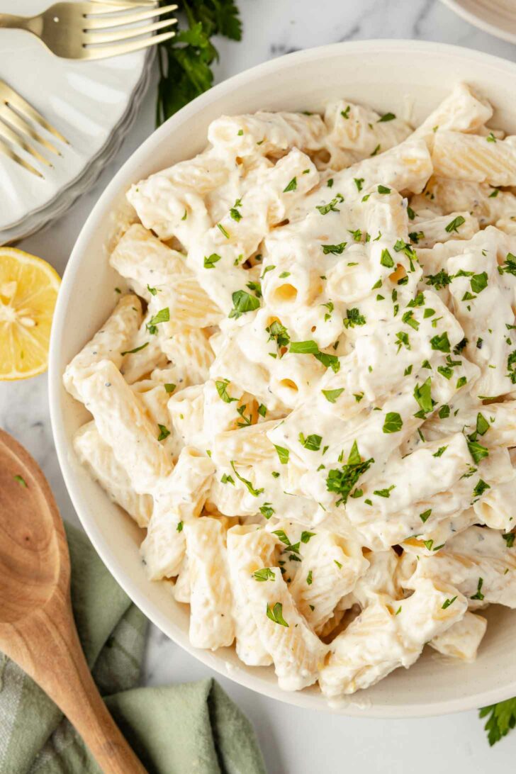 A bowl of rigatoni pasta covered in creamy white sauce, garnished with chopped parsley. A wooden spoon, lemon half, and forks are nearby on a marble surface.
