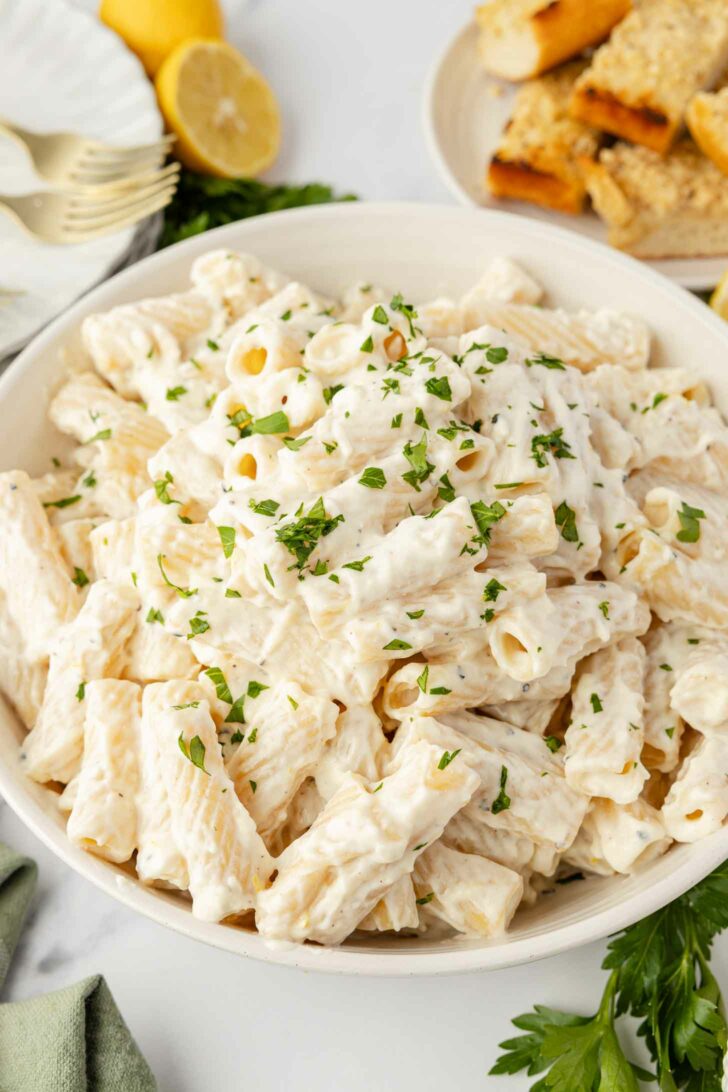 A bowl of rigatoni pasta coated in a creamy white sauce and garnished with chopped parsley, with breadsticks and lemon in the background.