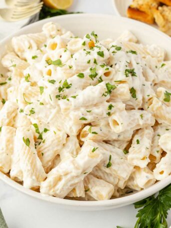 A large white bowl filled with creamy rigatoni pasta, garnished with chopped parsley. Slices of lemon, garlic bread, plates, and a wooden spoon are visible around the bowl on a white surface.