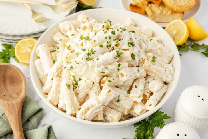 A large white bowl filled with creamy rigatoni pasta, garnished with chopped parsley. Slices of lemon, garlic bread, plates, and a wooden spoon are visible around the bowl on a white surface.