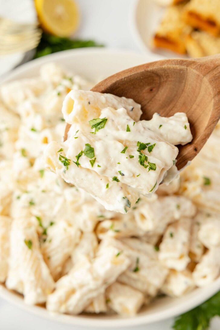 A wooden spoon holds a serving of creamy rigatoni pasta topped with chopped parsley, above a bowl filled with more of the same pasta. A blurred lemon wedge and bread are in the background.