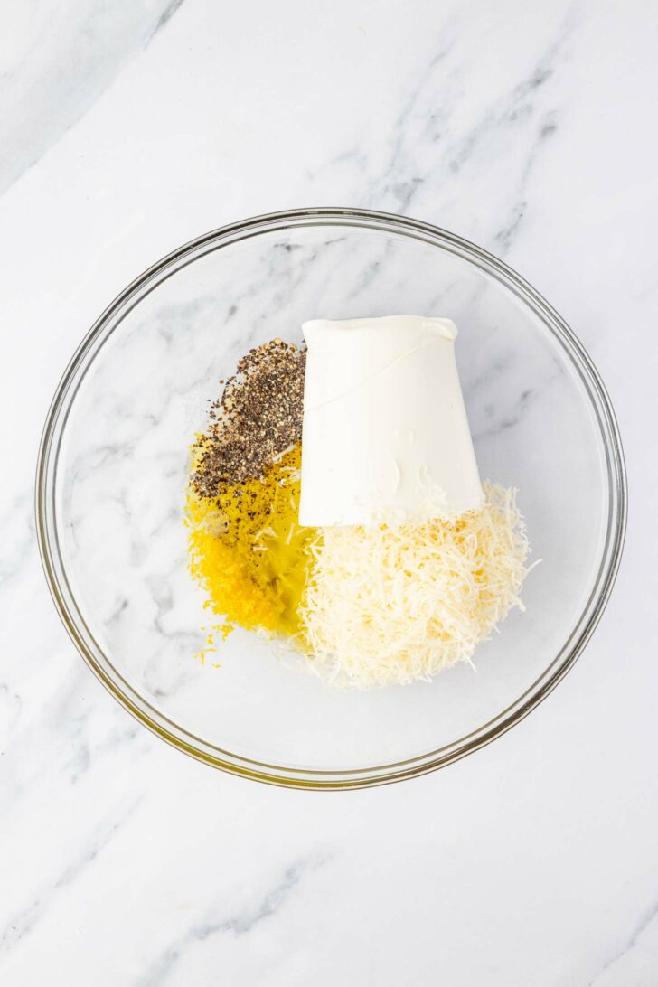 A glass bowl on a marble surface contains ricotta cheese, shredded parmesan cheese, lemon zest, and ground black pepper in separate piles.