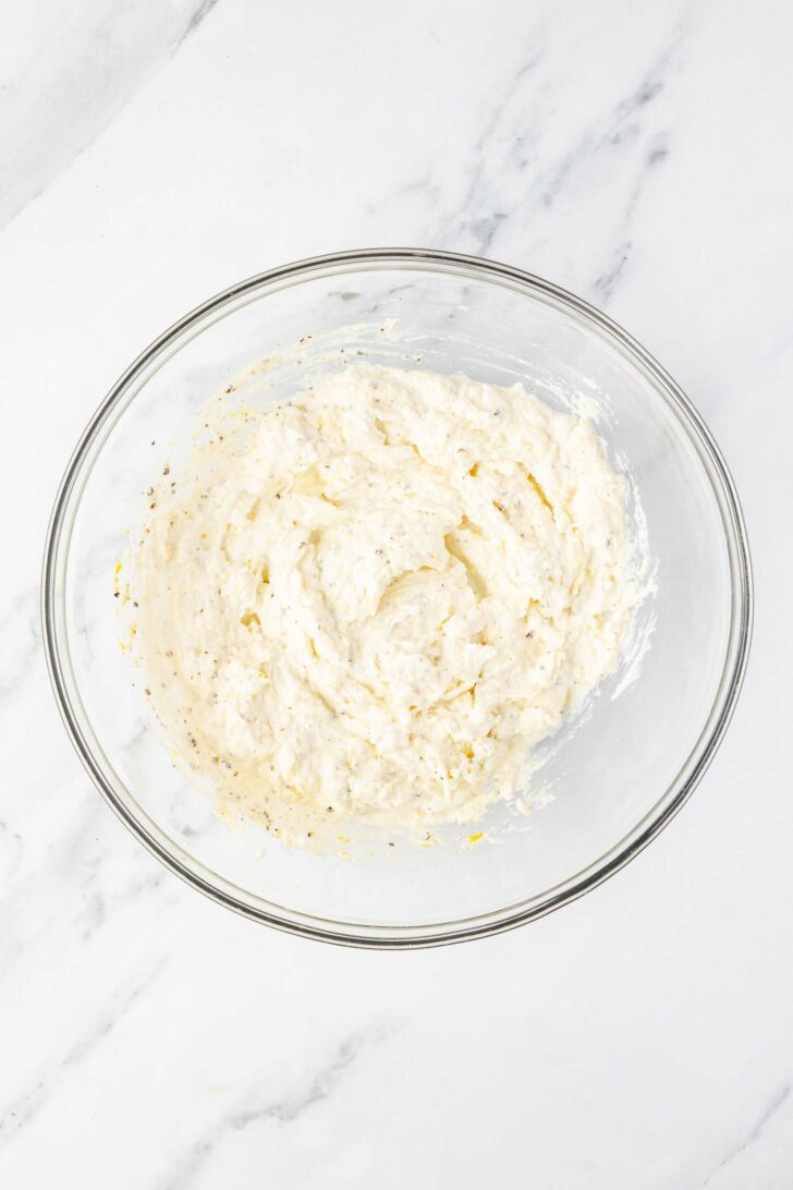 A clear glass mixing bowl filled with a creamy, white cheese mixture sits on a white marble countertop.