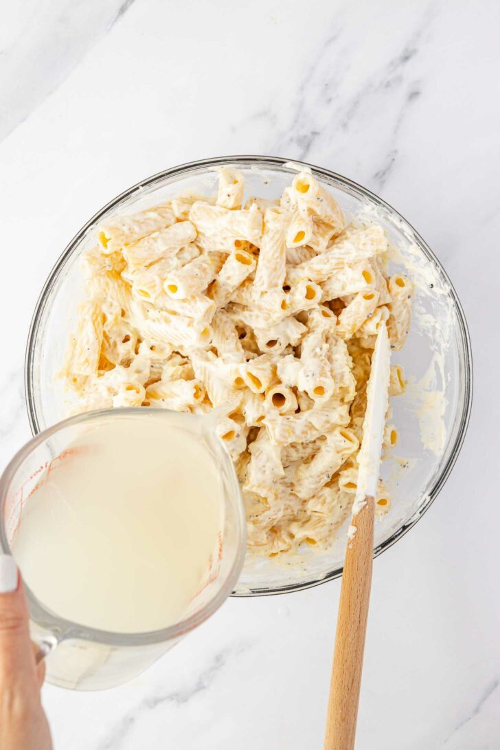 A glass bowl filled with cooked rigatoni pasta coated in a creamy white sauce sits on a marble surface. A hand is pouring a cup of starchy pasta water into the bowl, and a spatula rests inside.