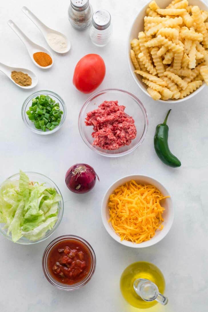 Assorted ingredients for nacho fries including crinkle-cut fries, ground beef, shredded cheddar, salsa, lettuce, green onion, red onion, tomato, jalapeño, oil, spices, and salt and pepper, arranged on a white surface.
