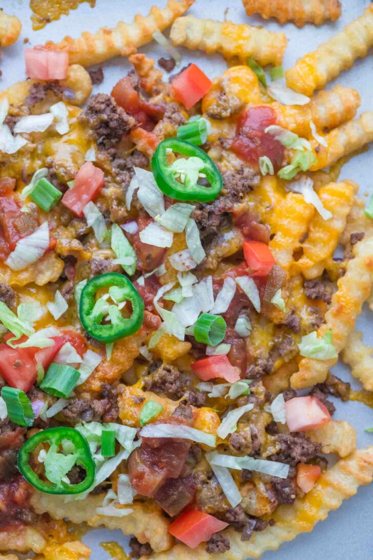 Close-up of crinkle-cut fries topped with melted cheese, ground beef, diced tomatoes, shredded lettuce, green onions, and sliced jalapeños.