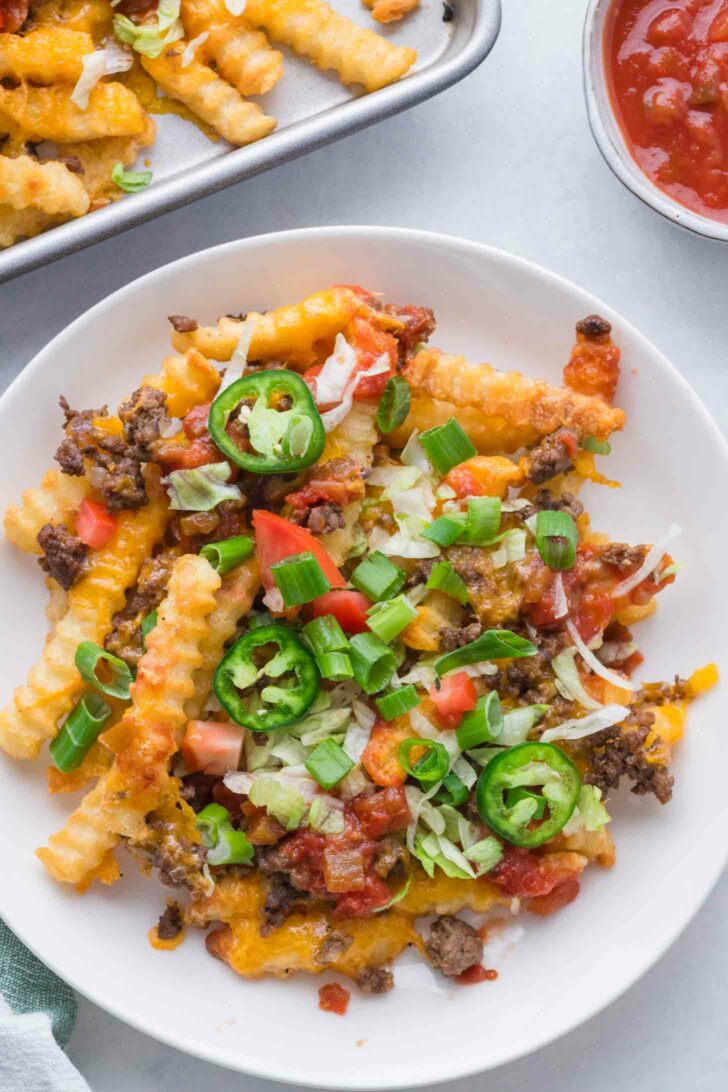 A plate of crinkle-cut fries topped with melted cheese, ground beef, diced tomatoes, shredded lettuce, sliced jalapeños, and chopped green onions, with salsa and more fries in the background.