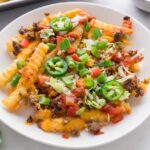 A plate of crinkle-cut fries topped with melted cheese, ground beef, chopped tomatoes, green onions, shredded lettuce, sliced jalapeños, and salsa. A green-and-white towel and a tray are visible nearby.