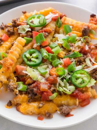 A plate of crinkle-cut fries topped with melted cheese, ground beef, chopped tomatoes, green onions, shredded lettuce, sliced jalapeños, and salsa. A green-and-white towel and a tray are visible nearby.