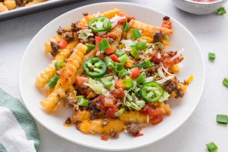 A plate of crinkle-cut fries topped with melted cheese, ground beef, chopped tomatoes, green onions, shredded lettuce, sliced jalapeños, and salsa. A green-and-white towel and a tray are visible nearby.