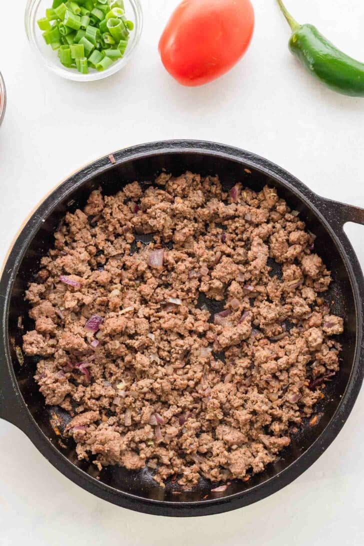 A black skillet filled with cooked ground beef sits on a white surface, surrounded by a bowl of chopped green onions, a whole tomato, and a green jalapeño pepper.