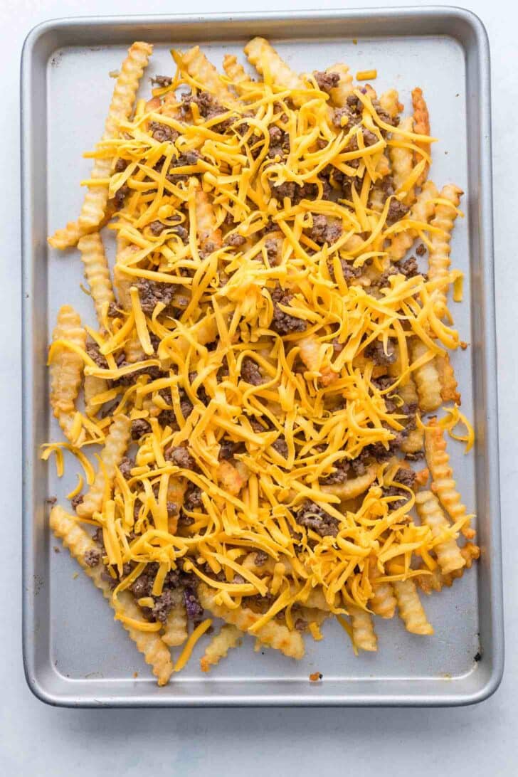 A baking tray filled with crinkle-cut French fries topped with ground beef and shredded cheddar cheese, ready to be baked.
